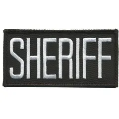 Sew-on embroidered insignia measuring 4 in x 2 in. Black and white design on plastic construction. Attaches to chest area via sewing. Used for uniform identification in security or tactical contexts.